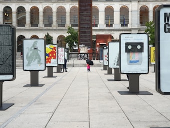 An outdoor exhibition displays various artistic posters on modern stands arranged in a plaza setting. A few people with umbrellas are seen walking between the displays. The background features an older building with arched windows, adding an architectural contrast to the modern art showcase.