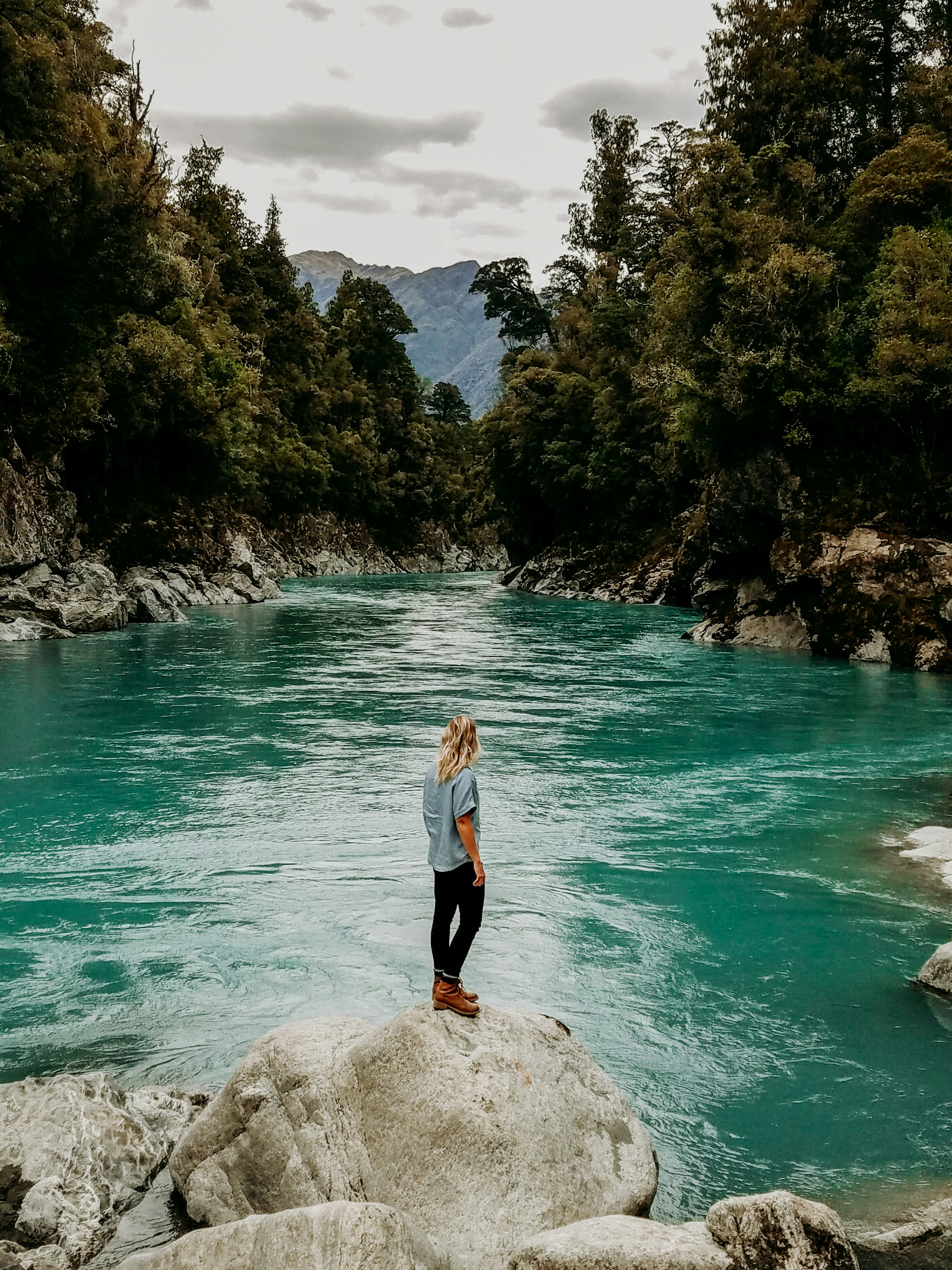 Blonde woman stands on rock overlooking Hokitika Gorge in New Zealand. 