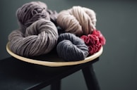 Bundles of yarn in various colors are arranged on a wooden plate. The yarn colors include shades of gray, beige, and dark pink, giving a cozy and soft texture. The background is dark, making the yarn colors stand out.