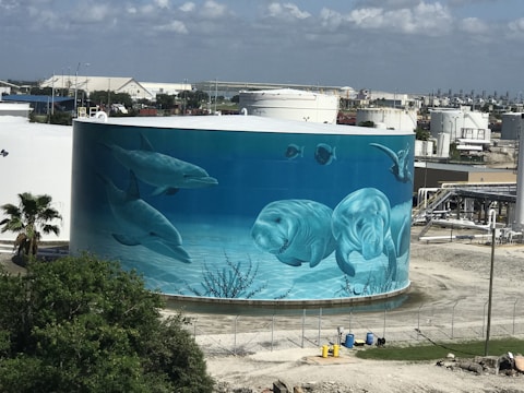A large cylindrical tank, likely a storage tank, features a vibrant mural depicting underwater marine life. The mural includes dolphins, manatees, and other aquatic creatures, painted in shades of blue to mimic an ocean scene. In the background, industrial buildings are visible, adding contrast between nature and industry.