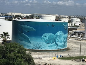 A large cylindrical tank, likely a storage tank, features a vibrant mural depicting underwater marine life. The mural includes dolphins, manatees, and other aquatic creatures, painted in shades of blue to mimic an ocean scene. In the background, industrial buildings are visible, adding contrast between nature and industry.