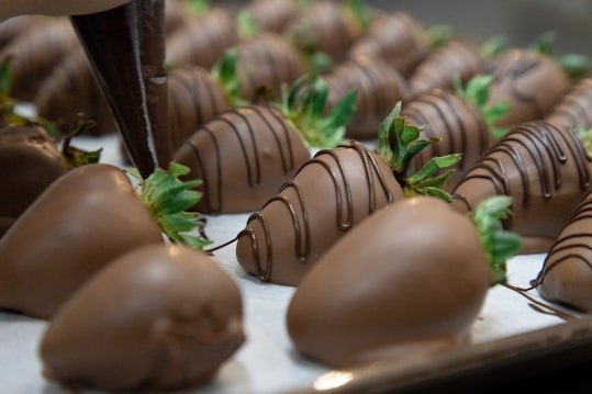 Numerous chocolate-covered strawberries are arranged on a tray, with a piping bag in the process of drizzling chocolate over some of them. The strawberries have green leaves and are uniformly coated in chocolate with a decorative drizzle on top.