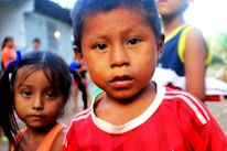 A young child wearing a red and white striped shirt is looking directly at the camera. Another child stands slightly behind, out of focus, with blue hair ties and an orange top. The background is blurred, featuring other children and a structure that resembles a house.
