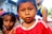 A young child wearing a red and white striped shirt is looking directly at the camera. Another child stands slightly behind, out of focus, with blue hair ties and an orange top. The background is blurred, featuring other children and a structure that resembles a house.