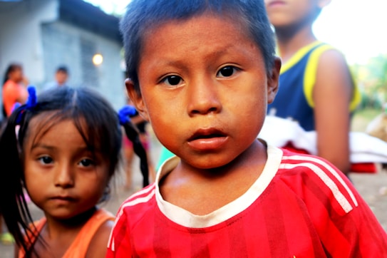 A young child wearing a red and white striped shirt is looking directly at the camera. Another child stands slightly behind, out of focus, with blue hair ties and an orange top. The background is blurred, featuring other children and a structure that resembles a house.