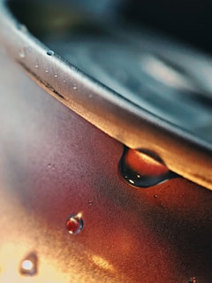 A close-up view of a metal can with condensation droplets forming on its surface. The image focuses on the top edge of the can, and the lighting highlights the droplets and creates a metallic sheen. The background is out of focus, emphasizing the water droplets and textures on the can.