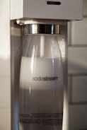 A close-up view of a Sodastream machine with a transparent bottle filled with carbonated water inside the machine. The Sodastream logo is visible on the bottle, and the machine has a white and metallic design.