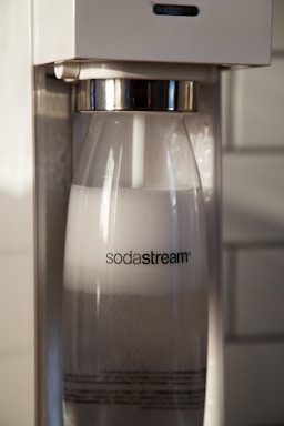 A close-up view of a Sodastream machine with a transparent bottle filled with carbonated water inside the machine. The Sodastream logo is visible on the bottle, and the machine has a white and metallic design.