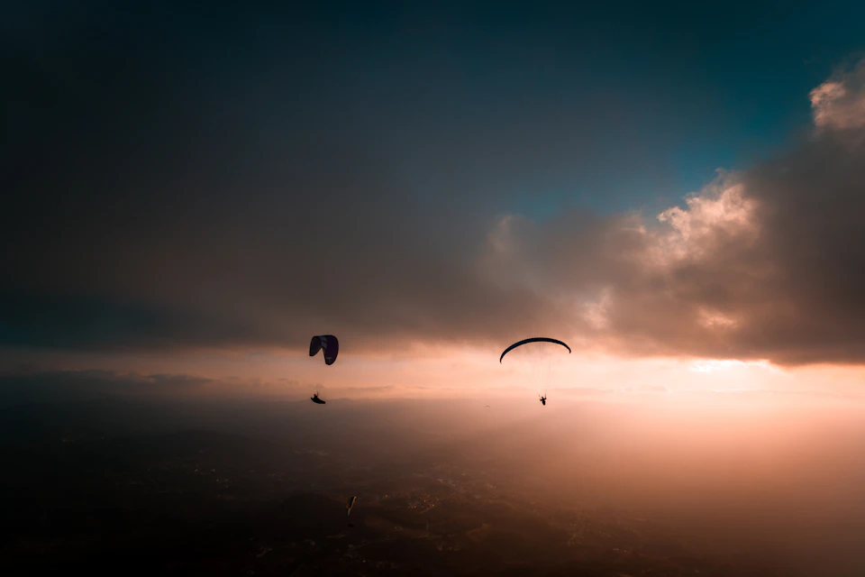 Two individuals paraglide against vibrant sunset over cityscape, contrasting sky and silhouetted figures