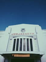 A large white building with an art deco design features a prominent clock at the top-center, with the word 'JOGJAKARTA' in red capital letters below it. A sign indicating 'Departure' is visible at the bottom, with text in both English and Indonesian.