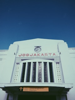 A large white building with an art deco design features a prominent clock at the top-center, with the word 'JOGJAKARTA' in red capital letters below it. A sign indicating 'Departure' is visible at the bottom, with text in both English and Indonesian.