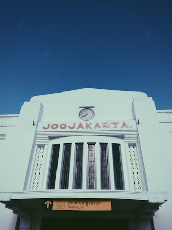 A large white building with an art deco design features a prominent clock at the top-center, with the word 'JOGJAKARTA' in red capital letters below it. A sign indicating 'Departure' is visible at the bottom, with text in both English and Indonesian.