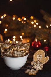 A bowl filled with festive, intricately decorated gingerbread cookies, featuring designs such as stars, mittens, and snowflakes. In the background, warm, blurred yellow lights create a cozy, holiday ambiance.