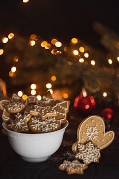A bowl filled with festive, intricately decorated gingerbread cookies, featuring designs such as stars, mittens, and snowflakes. In the background, warm, blurred yellow lights create a cozy, holiday ambiance.