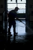A silhouette of a person wearing boots and a cap is using a hose to wash a floor inside a dimly lit space. The background features an ornate gate with a view of a bright outdoor area.