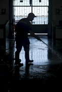 A silhouette of a person wearing boots and a cap is using a hose to wash a floor inside a dimly lit space. The background features an ornate gate with a view of a bright outdoor area.