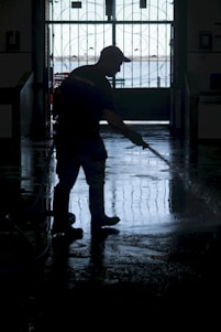 A silhouette of a person wearing boots and a cap is using a hose to wash a floor inside a dimly lit space. The background features an ornate gate with a view of a bright outdoor area.