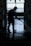 A silhouette of a person wearing boots and a cap is using a hose to wash a floor inside a dimly lit space. The background features an ornate gate with a view of a bright outdoor area.