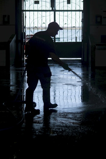 A silhouette of a person wearing boots and a cap is using a hose to wash a floor inside a dimly lit space. The background features an ornate gate with a view of a bright outdoor area.
