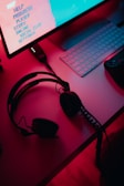 A dimly lit gaming setup with a monitor displaying a game's menu on the screen. A mechanical keyboard sits in front of the monitor. A pair of over-ear headphones and a game controller rest on the desk. The scene is bathed in a red and blue light, creating a moody gaming atmosphere.