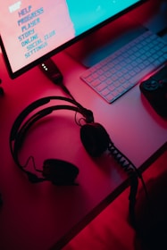 A dimly lit gaming setup with a monitor displaying a game's menu on the screen. A mechanical keyboard sits in front of the monitor. A pair of over-ear headphones and a game controller rest on the desk. The scene is bathed in a red and blue light, creating a moody gaming atmosphere.
