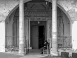 An elderly person sits alone on a chair in front of an old, weathered building with decorative elements above the doorway. Two large stone columns frame the entrance, and the overall look of the structure is dilapidated with peeling paint and rough surfaces. The mood is somber and solitary.