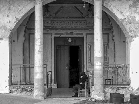 An elderly person sits alone on a chair in front of an old, weathered building with decorative elements above the doorway. Two large stone columns frame the entrance, and the overall look of the structure is dilapidated with peeling paint and rough surfaces. The mood is somber and solitary.