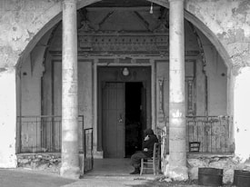 An elderly person sits alone on a chair in front of an old, weathered building with decorative elements above the doorway. Two large stone columns frame the entrance, and the overall look of the structure is dilapidated with peeling paint and rough surfaces. The mood is somber and solitary.