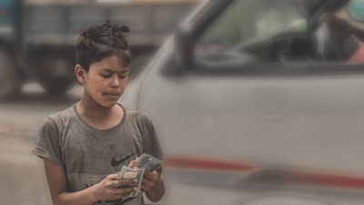 A young person stands outside, counting money with a focused expression. They are wearing a gray t-shirt and the background includes a blurred vehicle.