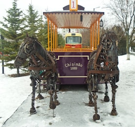 A tram with the label 'Chișinău 1889' is pulled by two life-sized horse sculptures made from metal and mechanical parts. The tram is painted in a rich purple color, with a bright red and yellow section in the back resembling a bus front. It is set in a snowy outdoor environment with several evergreen trees and a leafless tree in the background.