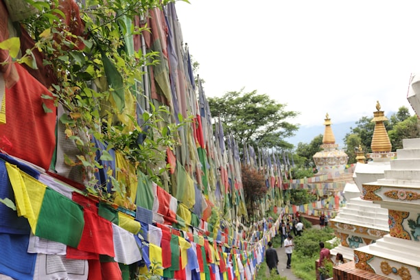 Colorful prayer flags are strung along a path surrounded by lush green trees and vegetation. The scene includes ornate, white stupas with golden tops, indicating a spiritual or religious site. Visitors can be seen walking along the path, adding a sense of scale and life to the tranquil setting.
