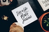 A tabletop features a motivational quote on a piece of paper that reads, 'Live a life that would make you jealous.' Accompanying the paper are various objects including a vintage pocket watch, a terracotta pot with a plant, and a couple of books, one of which is partially visible. The scene conveys a mix of retro and organic elements.