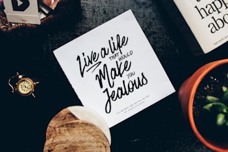 A tabletop features a motivational quote on a piece of paper that reads, 'Live a life that would make you jealous.' Accompanying the paper are various objects including a vintage pocket watch, a terracotta pot with a plant, and a couple of books, one of which is partially visible. The scene conveys a mix of retro and organic elements.