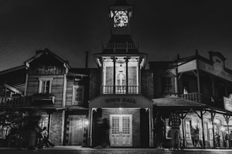 A nighttime scene depicting an old-style town hall with classic Western architecture, featuring wooden structures and a prominent clock tower. The building facade is illuminated by soft lighting, creating dramatic shadows. Several figures are present, adding a sense of liveliness to the scene.