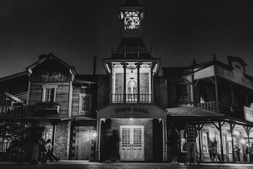 A nighttime scene depicting an old-style town hall with classic Western architecture, featuring wooden structures and a prominent clock tower. The building facade is illuminated by soft lighting, creating dramatic shadows. Several figures are present, adding a sense of liveliness to the scene.