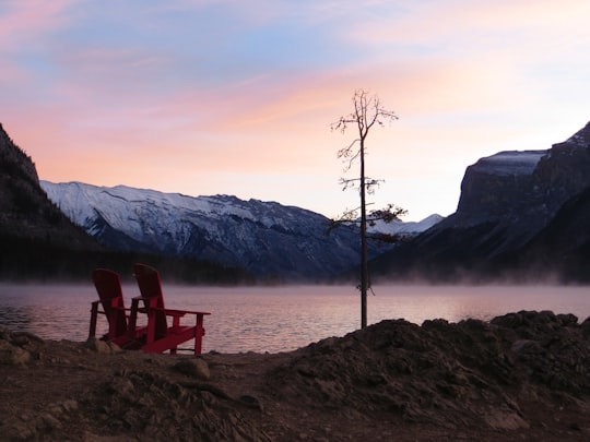 two red wooden adirondack chairs beside water in Lake Minnewanka Canada
