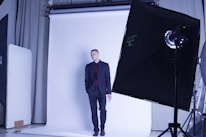 A person in a suit stands against a white backdrop in a photography studio. Professional lighting equipment, including softboxes and reflectors, is visible.