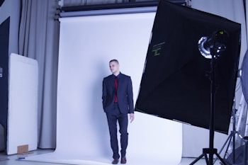 A person in a suit stands against a white backdrop in a photography studio. Professional lighting equipment, including softboxes and reflectors, is visible.