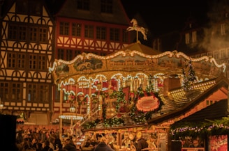 A vibrant and festive scene of a traditional Christmas market with a vintage carousel lit up at night. The carousel is adorned with lights and festive decorations, including garlands and ornaments. In the background, there are timber-framed buildings typical of European architecture. The market is bustling with people enjoying the festive atmosphere.