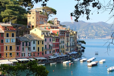 A scenic coastal village with vibrant, colorful buildings featuring Italian architecture lining a calm waterfront. Small boats are moored in the tranquil water, and lush hills surround the area.