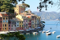 A scenic coastal village with vibrant, colorful buildings featuring Italian architecture lining a calm waterfront. Small boats are moored in the tranquil water, and lush hills surround the area.