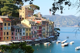 A scenic coastal village with vibrant, colorful buildings featuring Italian architecture lining a calm waterfront. Small boats are moored in the tranquil water, and lush hills surround the area.