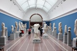 A spacious museum gallery with high arched ceilings and walls adorned in blue showcases a collection of classical marble statues and busts. Visitors are scattered throughout, some taking notes and others observing the artwork closely. The sculptures are displayed on pedestals aligned in rows along the room.