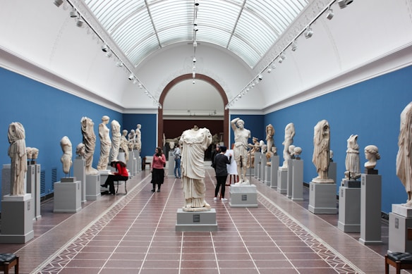 A spacious museum gallery with high arched ceilings and walls adorned in blue showcases a collection of classical marble statues and busts. Visitors are scattered throughout, some taking notes and others observing the artwork closely. The sculptures are displayed on pedestals aligned in rows along the room.