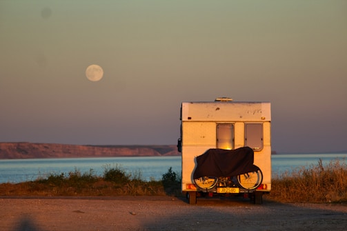 A campervan is parked near a coastal area with a bicycle attached to its rear. The sea is visible in the background, and a full moon hangs in the sky. The landscape is serene with grassy patches and a distant rocky shoreline.