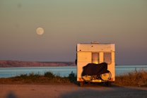 A campervan is parked near a coastal area with a bicycle attached to its rear. The sea is visible in the background, and a full moon hangs in the sky. The landscape is serene with grassy patches and a distant rocky shoreline.