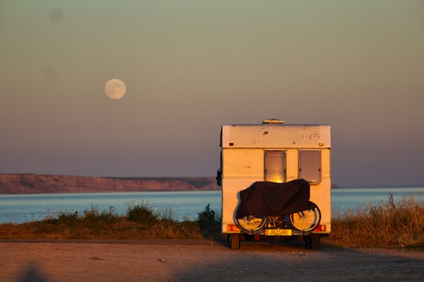 A campervan is parked near a coastal area with a bicycle attached to its rear. The sea is visible in the background, and a full moon hangs in the sky. The landscape is serene with grassy patches and a distant rocky shoreline.