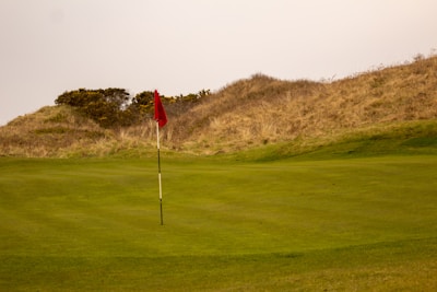 A grassy golf course with a red flag on a pole marking the hole. Surrounding the green are gentle hills covered in brownish grass and some bushes.