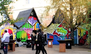 A vibrant and colorful mural covers the walls of small, house-shaped structures. The artwork features comic book graphics, including fists, speech bubbles with the word 'boom', and vivid, stylized images of superheroes. Several people in winter clothing are gathered around the area, appearing to enjoy the scene. The setting is partially surrounded by trees showing autumn foliage.