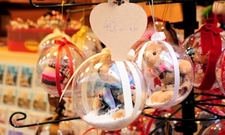 Holiday-themed ornaments displayed in a market. Each ornament contains a small teddy bear and artificial snow inside a clear plastic sphere. They are adorned with colorful ribbons, and a heart-shaped tag with pricing information is attached to one of the ornaments.